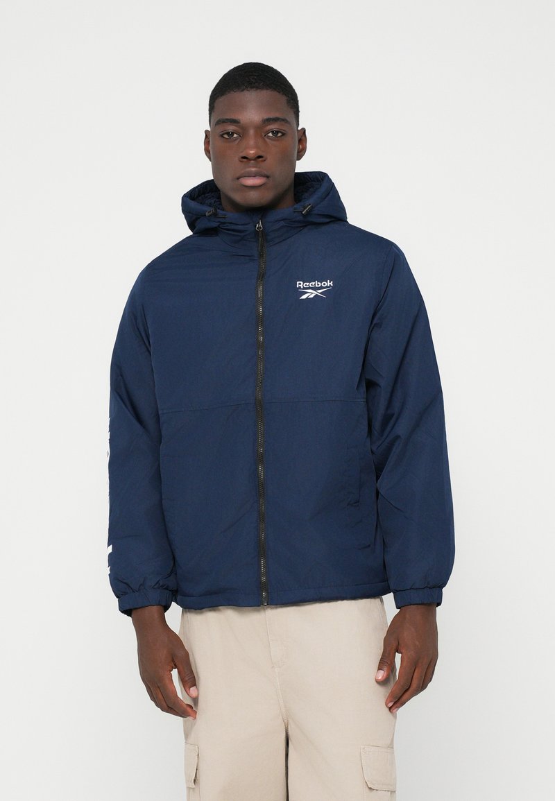 Tânăr îmbrăcat cu o jachetă cu glugă Reebok de culoare bleumarin, cu fermoar în față, și pantaloni cargo bej, stând în fața unui fundal alb simplu.