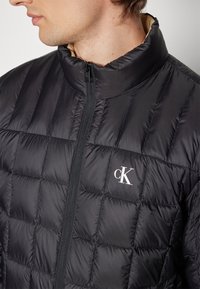 Doudoune noire matelassée avec un col haut, une fermeture éclair et un logo "CK" blanc sur la poitrine. Texture de tissu lisse et brillant.