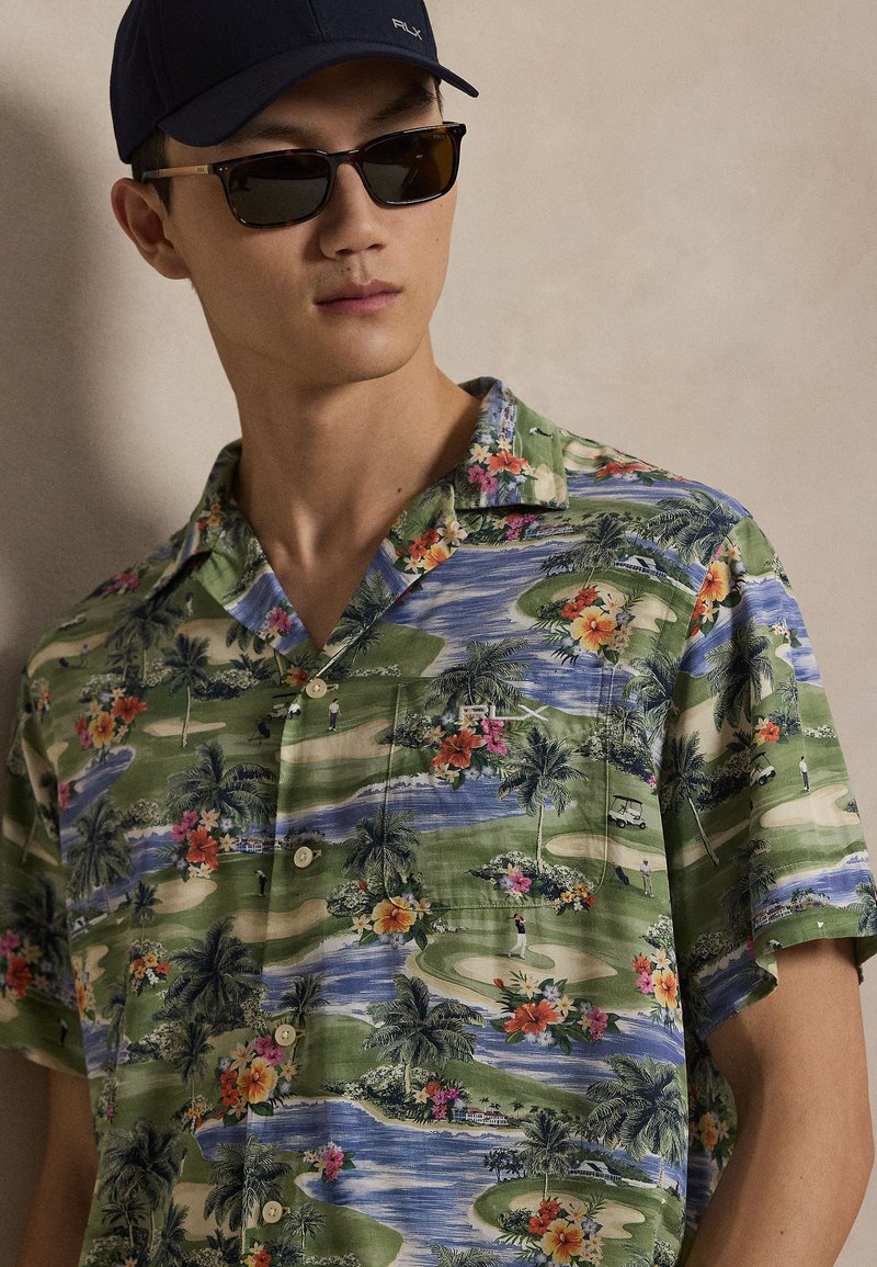 Short-sleeve button-up shirt in a green tropical print featuring flowers, palm trees, and a golf course design. Made of lightweight fabric.