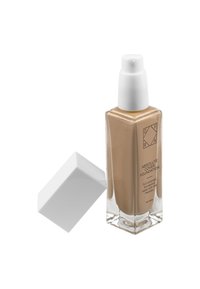 Bottiglia di fondotinta in vetro trasparente con bordi angolati, riempita con un liquido color tan, con un erogatore a pompa bianco e un coperchio quadrato.