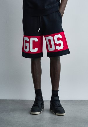 Άνδρας που φοράει μαύρο και κόκκινο σορτς με μεγάλες λευκές γράμματα "GCDS", μαύρες κάλτσες και μαύρα αθλητικά παπούτσια, στέκεται μπροστά σε ένα απλό γκρι φόντο.
