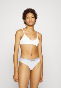 Hvit ribbet bralette med tynne stropper, kombinert med hvite bikini-lignende undertøy med en grå logo på livet. Glatt stofftekstur.
