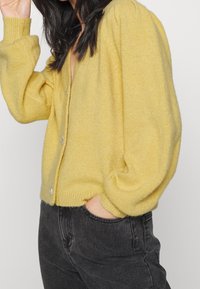 Žena v žlutém button-up kardiganu s napušťenými rukávy a černými džínami, s jednou rukou v kapse, otočená na stranu proti jednoduchému pozadí.