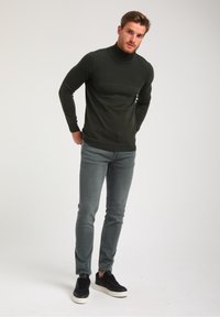 Dunkelgrüner, langärmeliger Rollkragenpullover mit strukturiertem Oberteil, kombiniert mit engen grauen Jeans und schwarzen Sneakers, die vor einem hellen Hintergrund stehen.