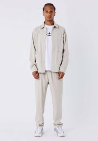 Lichtgrijs, gestructureerd stoffen shirt en bijpassende broekenset. Het shirt heeft een kraag, zijzakken en een knoopsluiting. Witte t-shirt eronder.