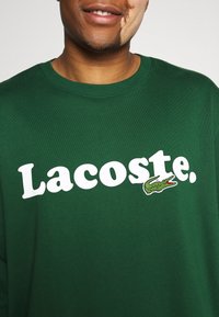 Felpa verde in cotone con grande logo bianco "Lacoste" e logo coccodrillo ricamato con dettagli rossi. Collo rotondo, vestibilità regolare.