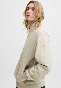 Veste bomber beige avec un col montant, des poignets côtelés, des poches zippées et un patch en tissu avec un logo sur la manche gauche.