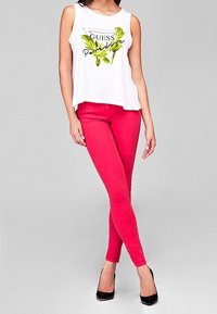 Biała bezrękawnik z kwiatowym nadrukiem i tekstem, zestawiony z jaskrawoczerwonymi skinny jeans i czarnymi szpilkami, stojący na szarym tle.