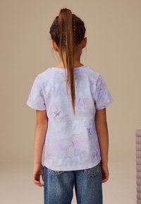 Tricou liliac cu mâneci scurte și imprimeu cu fluture, purtat cu blugi denim albaștri. Tricoul are un decolteu rotund și o croială lejeră.