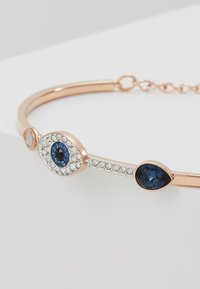 Rose gold bangle bracelet featuring an eye design with blue and clear stones, including one central round stone, and a sleek, smooth finish.