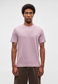 Άνδρας με κοντά σγουρά μαλλιά στέκεται στραμμένος προς τα εμπρός φορώντας ανοιχτό μωβ μπλουζάκι και σκούρα καφέ παντελόνια σε ουδέτερο φόντο.