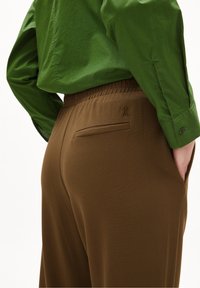 Brązowe spodnie z teksturowanym wykończeniem i elastycznym paskiem, z jedną tylną kieszenią oraz subtelnym haftem. Widoczna zielona koszula.