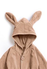 Tan färgad jacka med huva och kaninöron samt en enkel knappstängning. Mjuk textur, raglanärmar och en mysig design.