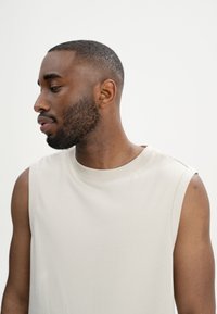 Hellbeige ärmelloses T-Shirt mit Rundhalsausschnitt. Glattes Baumwollmaterial und lockere Passform. Minimale Nähte an den Schultern.