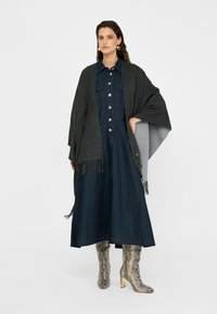 Donkergrijze poncho met franjes die over een blauwe denim jurk met knopen en zakken is gedrapeerd. Knielange laarzen met slangenprint maken de look compleet.