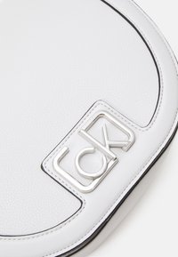 Weiße runde Tasche aus strukturiertem Leder, mit einem silbernen Logoakzent und schwarzem Stickdetail am Rand.