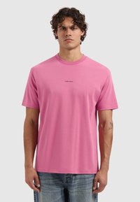 Mann mit lockigem Haar, der ein pinkes T-Shirt mit der Aufschrift "PURE PATH" und blaue Jeans trägt, steht vor einem schlichten hellgrauen Hintergrund.