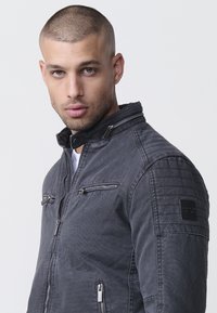 Sötétszürke farmerdzseki, bordázott vállrész kialakítással, cipzáras zsebekkel és patentos, magas gallérral. Észlelhető texturált anyag részlet.