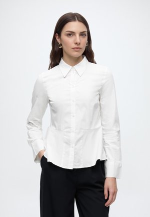 Woman with long brown hair wearing gold earrings, a white buttoned shirt, and black pants, standing with one hand in pocket.