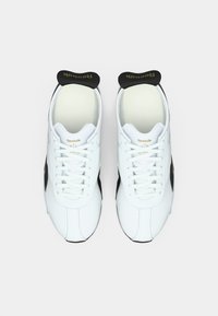 O pereche de tenisi Reebok albi cu tab-uri negre la călcâi și șireturi, văzuți de sus pe un fundal alb simplu.