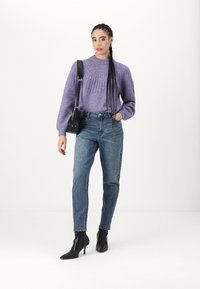 Pull en tricot violet avec texture côtelée, jeans en denim bleu et bottines noires à la cheville. Le mannequin tient un petit sac à main noir.