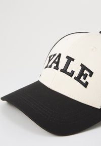 Beige színű korona fekete hímzéssel, amelyen a "YALE" felirat olvasható, és kontrasztos fekete napellenző. Tartós anyagból készült, strukturált dizájnnal.