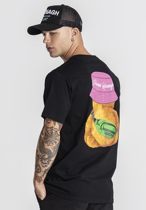 Jeune homme au bras tatoué portant une casquette noire et un t-shirt noir représentant un ours en peluche avec un chapeau rose et une sacoche verte dans le dos.