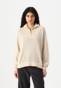 Beige, strikket genser med kvart glidelås, ribbestrikket krage og løs passform. Har åpen vev tekstur og droppe skuldre. Kombinert med mørke bukser.