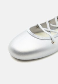 Ασημένιο μπαλαρίνα με ελαστικούς λουρίδες που διασταυρώνονται και στρογγυλεμένη μύτη, παρουσιασμένο σε λευκό φόντο.