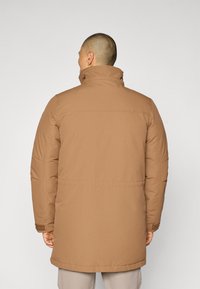 Brun polstret frakk med høy krage og sidelommer. Stoffet ser glatt ut, med rett snitt og midt-lår lengde.