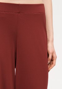 Κοντινό πλάνο ενός ατόμου που φοράει φαρδιά παντελόνια σε σκουριά χρώμα με ψηλή μέση και επίπεδη ζώνη, εμφανίζοντας μέρος του δεξιού του χεριού.