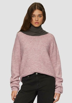 Roze gebreide trui met een bootnek en verlaagde schouders, gecombineerd met een donkergrijze coltrui eronder. Zwarte jeans worden gedragen.