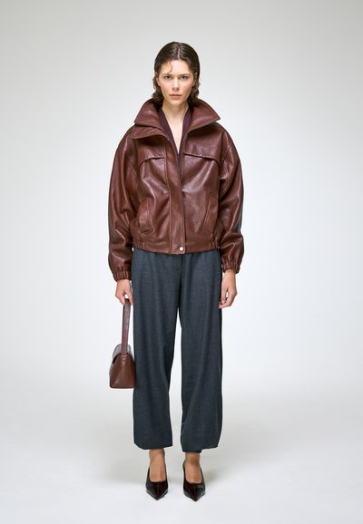 Femme portant une veste en cuir marron, un pantalon large gris, tenant un sac à main marron, et des talons pointus noirs sur un fond gris uni.
