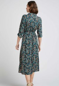 Robe midi florale avec une base en bleu sarcelle, des motifs multicolores et une taille cintrée. Manches longues avec poignets retroussés, tissu léger.
