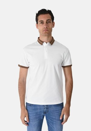 Uomo che indossa una polo bianca con colletto e bordi delle maniche marroni, abbinata a jeans blu, in piedi davanti a uno sfondo semplice.