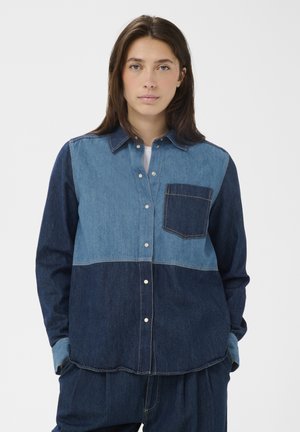 Femme portant une chemise en denim bicolore avec fermeture à boutons et poche poitrine, debout les mains dans les poches du pantalon, sur un fond uni.