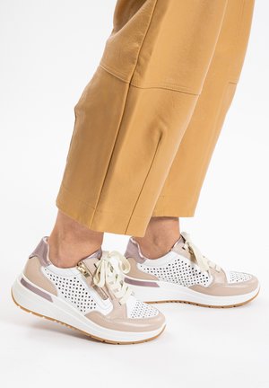 Person trägt beige, gekürzte Hosen und weiße-beige perforierte Sneakers mit Schnürsenkeln und seitlichem Reißverschluss, steht vor weißem Hintergrund.