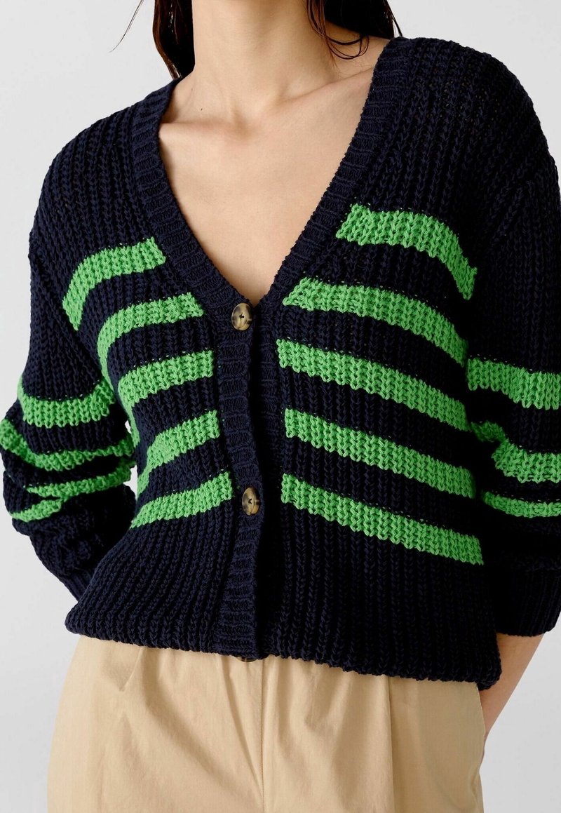 Námořnicky modrý pletený cardigan s zelenými pruhy, výstřihem do V a dvěma mosaznými knoflíky vpředu. Měkká textura s volným střihem.