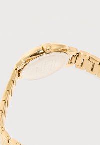 Μεταλλικό λουρί χειρολογίου με χρυσό τόνο και πλάγια όψη στρογγυλής θήκης ρολογιού με κορόνα, που δείχνει χαραγμένες λεπτομέρειες στην πίσω πλάκα, σε λευκό φόντο.