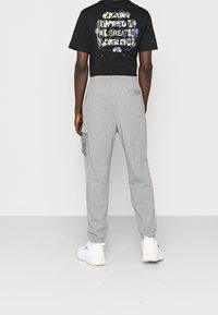 Schwarzes Crop-Top mit buntem Grafikdruck auf dem Rücken, kombiniert mit grauen Jogginghosen mit Cargo-Tasche und Netzdetails. Weiße Sneaker.