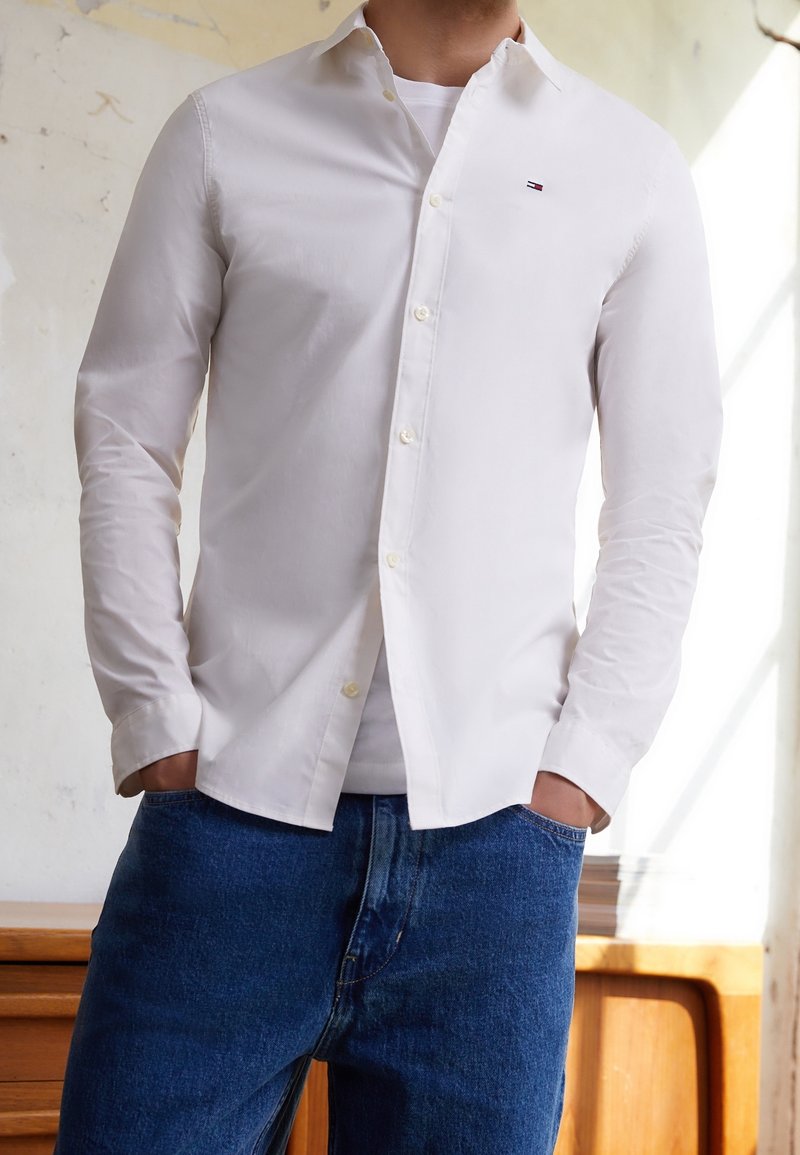 Uomo che indossa una camicia bianca con bottoni sopra una maglietta bianca e jeans blu, in piedi con le mani nelle tasche all'interno.