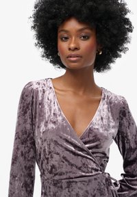 Fioletowa welurowa sukienka o długości, z długimi rękawami, głębokim dekoltem w kształcie litery V oraz z detalem w postaci związania w talii, charakteryzująca się miękką, teksturowaną powierzchnią i subtelnym blaskiem.