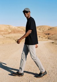 Homme portant une chemise noire, une casquette grise, des lunettes de soleil et un pantalon gris clair marchant sur un chemin poussiéreux dans un paysage désertique sableux et vallonné.