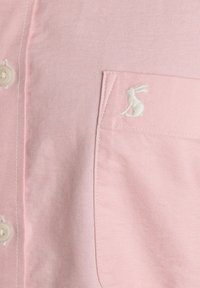 Chemise boutonnée rose clair avec une poche avant ornée d'un petit logo brodé d'un lapin blanc dans le coin supérieur.