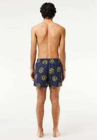 Bañador de marina con cinturilla elástica, con logotipos circulares verdes en una tela suave. La vista trasera muestra el corte relajado y una longitud por encima de la rodilla.
