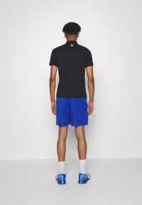 En man står vänd bort med en svart pikétröja, blå shorts, vita strumpor och blå atletiska skor mot en enkel vit bakgrund.