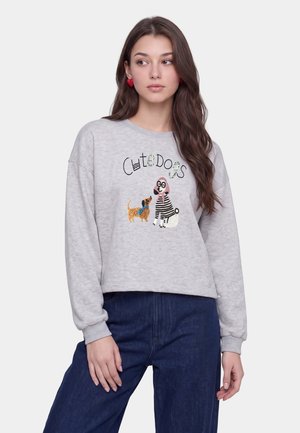Jonge vrouw draagt een lichtgrijze trui met de tekst "Cute Dogs" en twee cartoonhonden, gecombineerd met donkerblauwe jeans en rode hartvormige oorbellen.