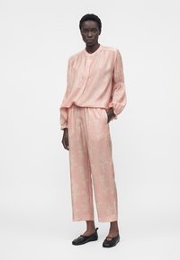 Vrouw die een lichtroze blouse met lange mouwen en bijpassende wijd uitlopende broek draagt met een subtiel bloemmotief, gecombineerd met zwarte platte schoenen.