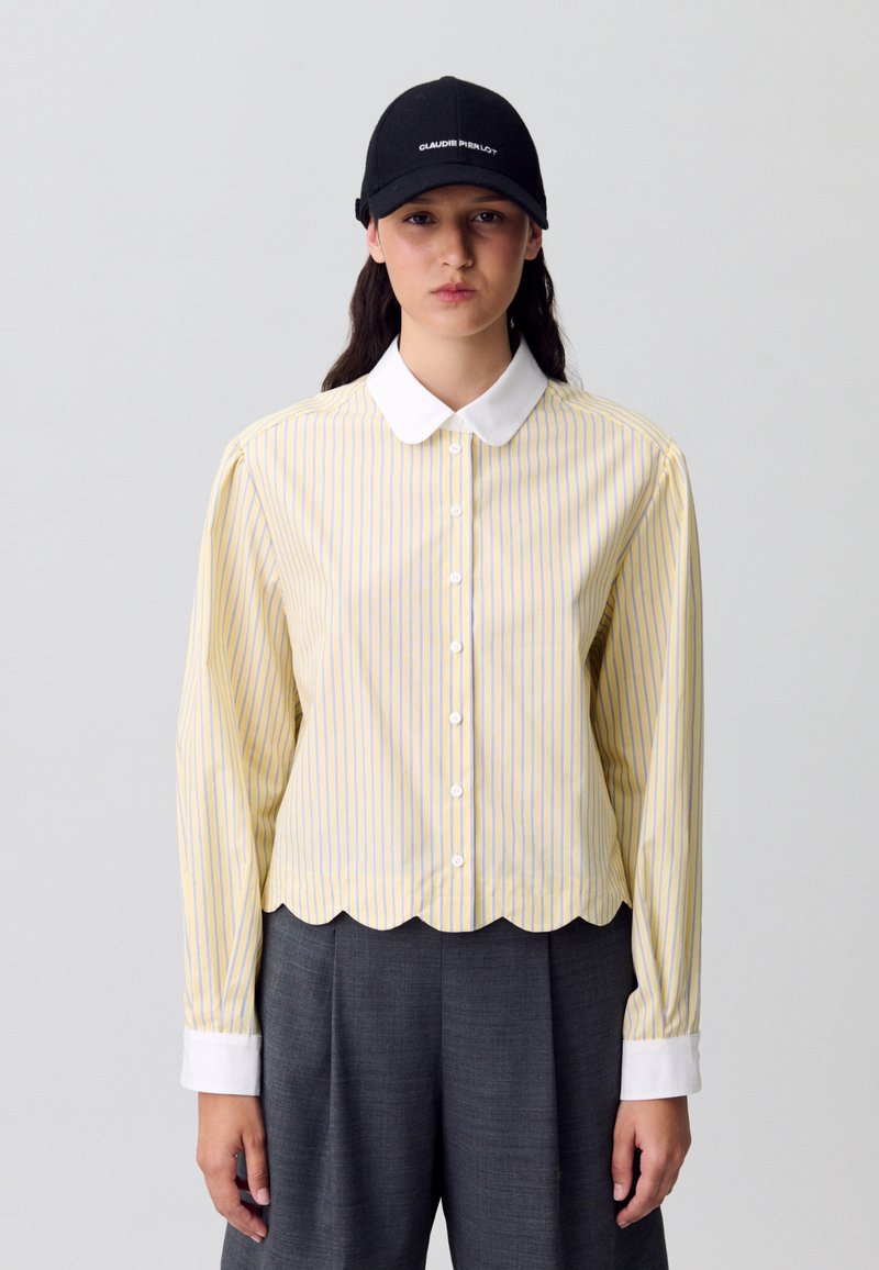 Camicia a righe gialla e blu con colletto bianco e orlo scallop, abbinata a pantaloni grigi a gamba larga e un berretto nero.