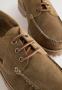 Braune Wildlederschuhe haben eine abgerundete Zehe, Leder-Schnürsenkel und kontrastierende weiße Nähte entlang der Nähte und der Sohle für zusätzliche Details.